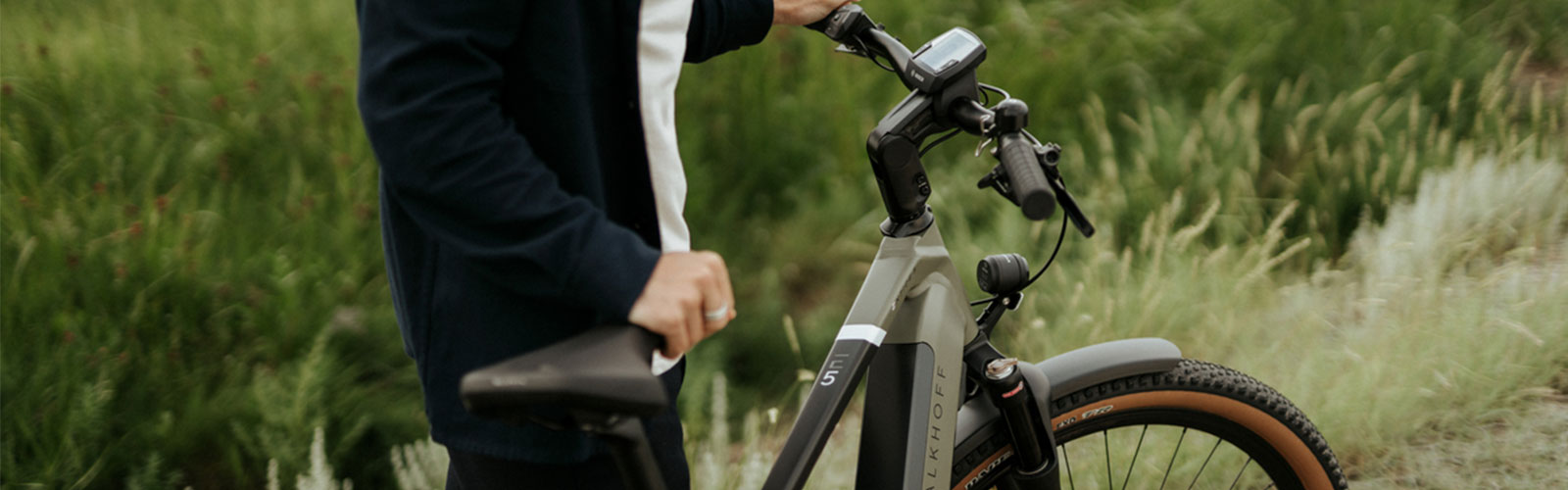Person schiebt ein Trekkingbike mit breiten Reifen durch eine grüne Wiesenlandschaft.
